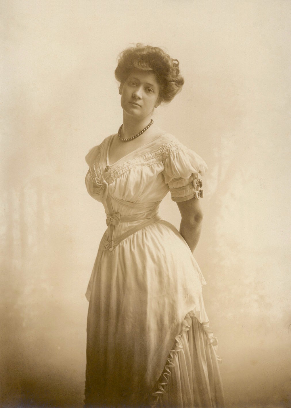 A beautiful image of Miss Ima Hogg in classic sepia hues. She has on a light colored 1900's style dress with her dark hair up in the fashion of the day. She looks to be in her 20s.